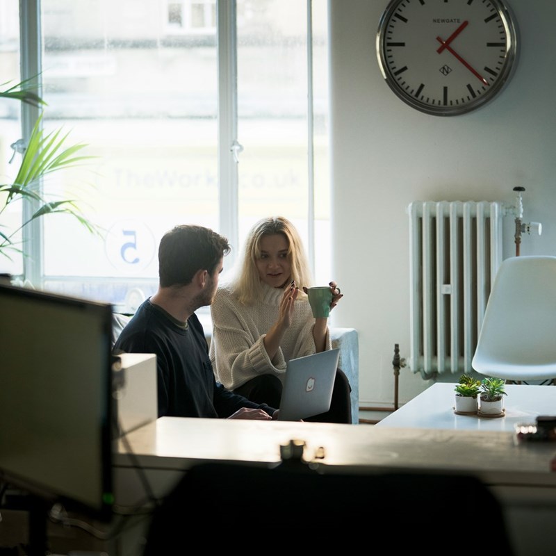 two people sitting on a couch talking, one holding a mug while using a laptop in a bright room.