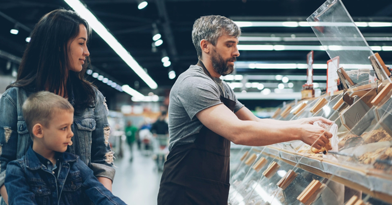 A male supermarket employee stocks bakery shelves as a mom and her young son watch