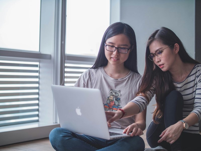 Two casually dressed women sit on the floor and look at a computer.