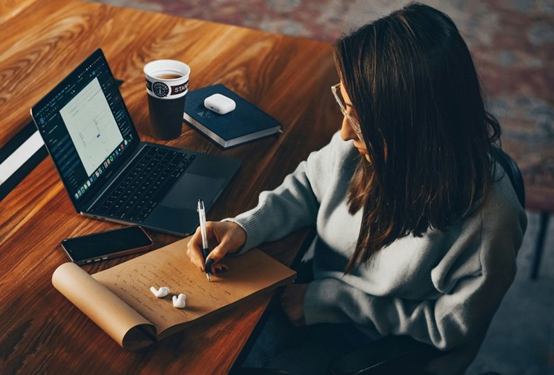 Woman wearing a grey sweatshirt, sitting down in a table while writing on paper and working with her computer.