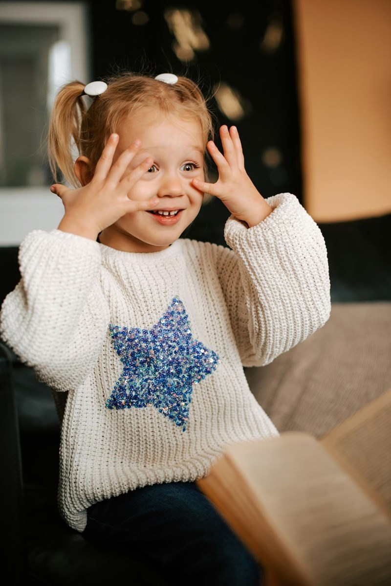 Little girl with pigtails and a sweatshirt with a sparkling star on it holds her hands up to her face and smiles