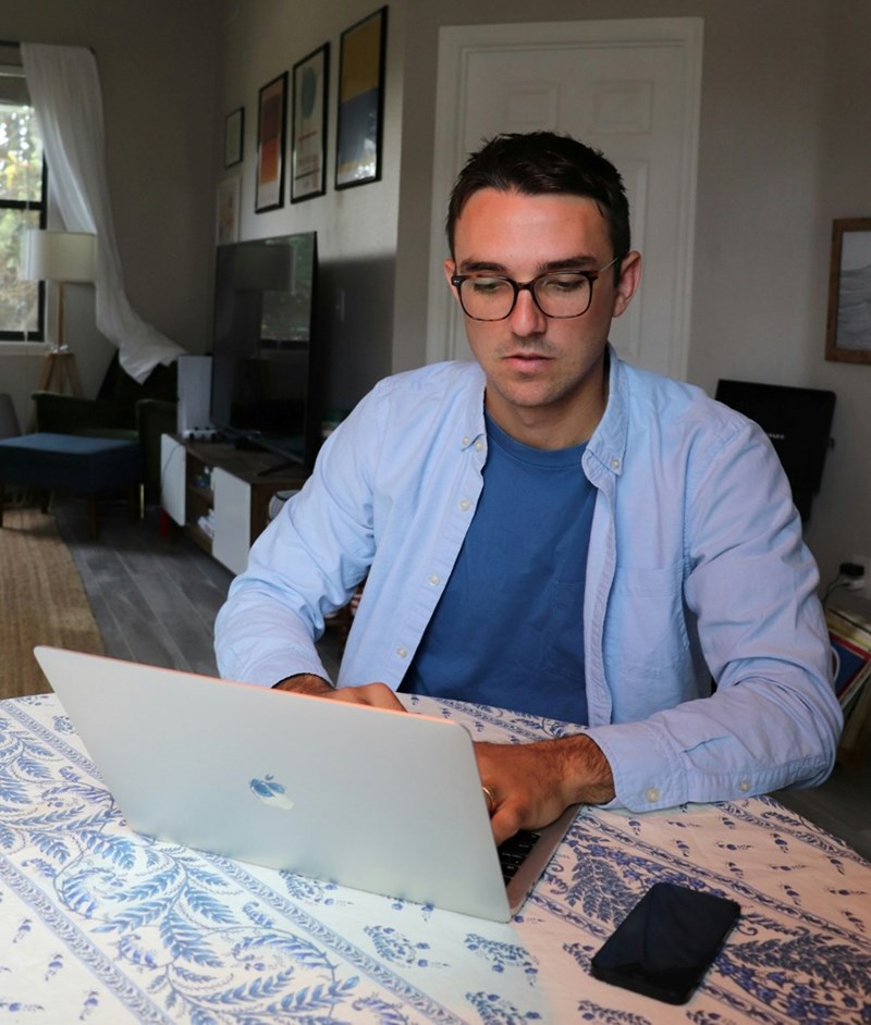 A man works on his laptop in his living room, his cell phone beside him on the table