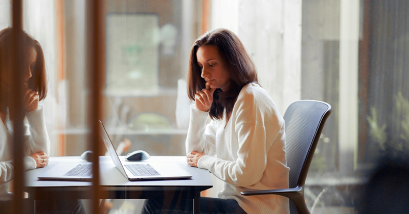 A female employee and her reflection, thinking as she works on her laptop in a meeting room