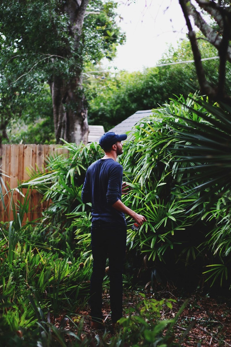 Man in backyard garden looking at dense greenery while holding small tool among plants.