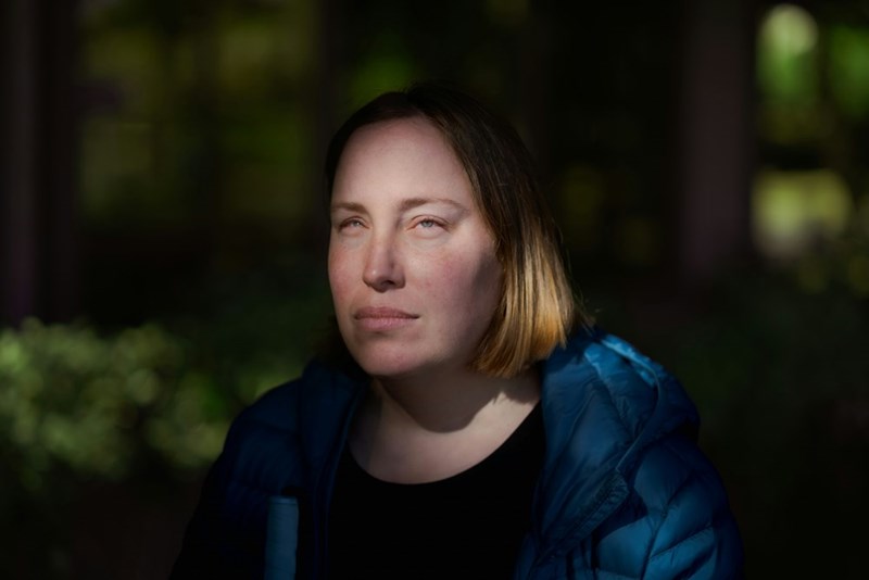 An unhappy woman in a blue jacket gives an angry stare while standing in front of a large plant.