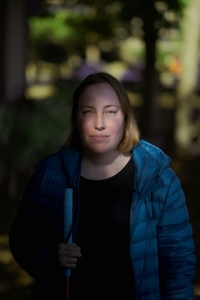 An unhappy woman in a blue jacket is obscured by a shadow as she stands outside.