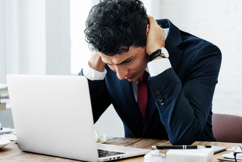 A frustrated employee puts his hands on the back of his neck while sitting in front of his laptop.