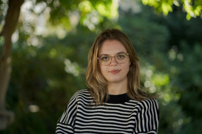 Woman with glasses in striped sweater looking at camera outdoors with blurred green background.