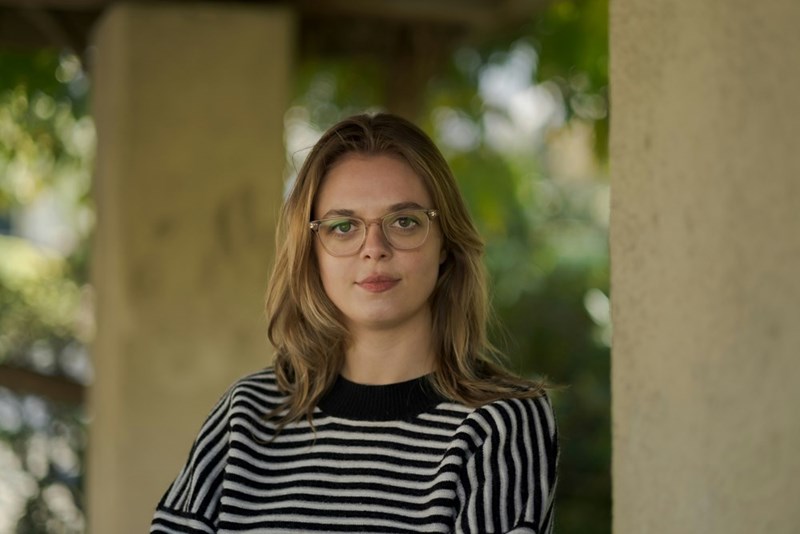 Woman in striped sweater and glasses standing between columns outdoors, looking at camera.
