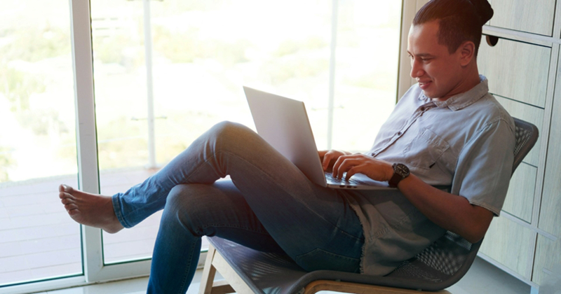 A shoeless man leans back in his chair as he works on a laptop at home