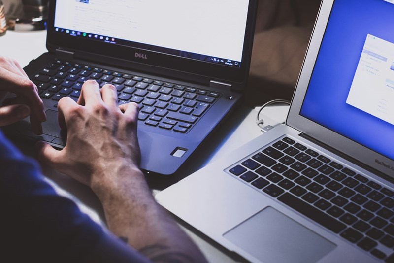 A man types on a black laptop next to another computer at his desk.