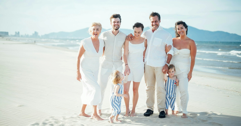 Five adults and two young children in a family pose together on a sunny beach