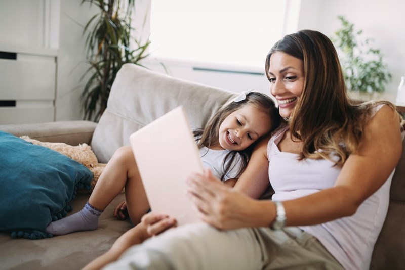 Mom and daughter smiling while using tablet together on couch at home