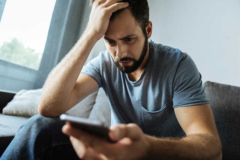 Frustrated man sitting on a grey couch looking at his phone while holding his head.