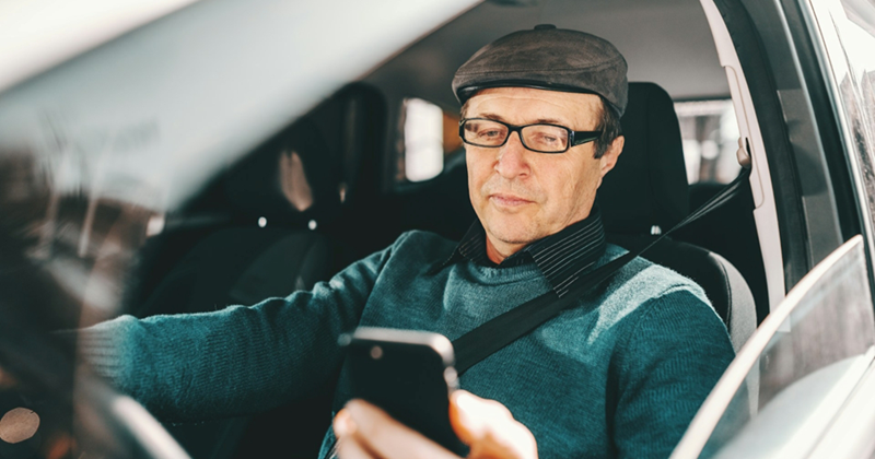 An older man strapped into the driver's seat of a car looks at his cell phone