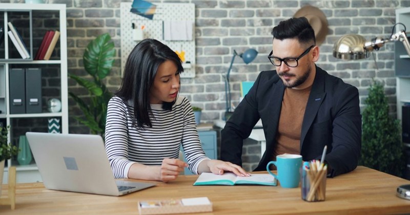A male and female coworker sit next to one another at a table looking at a laptop.