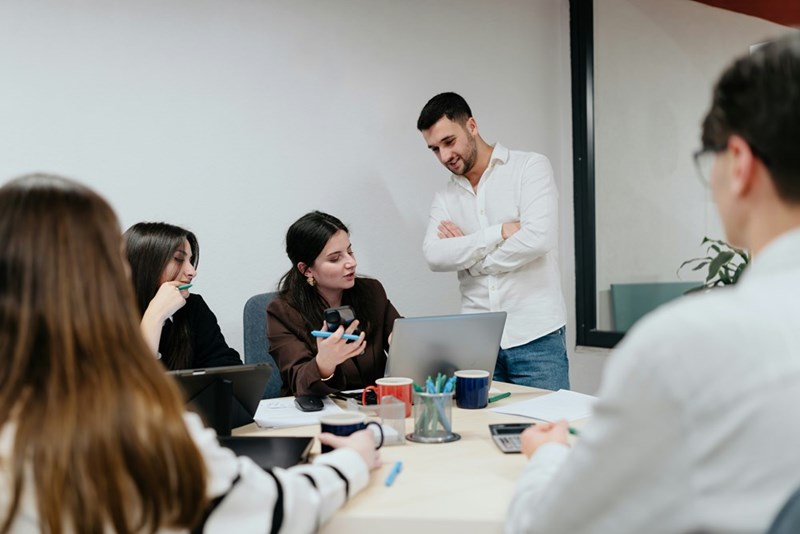 A manager looks over a team member's shoulder with folded arms as she explains something in a team meeting.