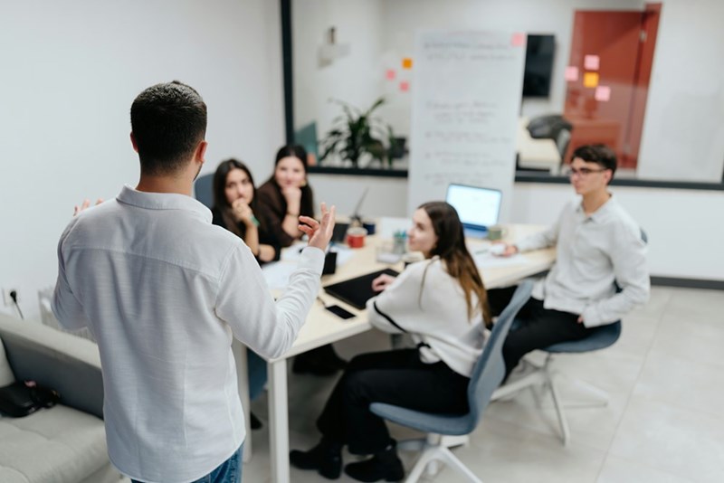 A manager addresses his team with his back to the camera in a conference room.