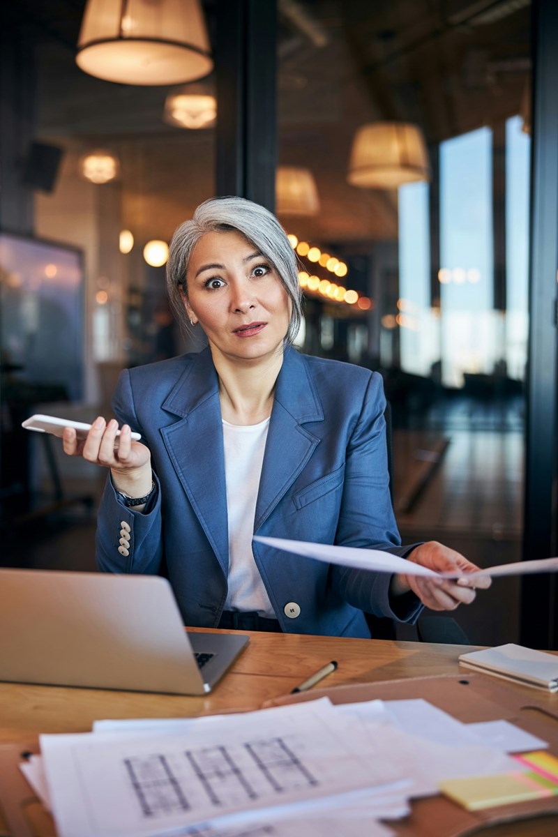 Puzzled woman holding smartphone and document while sitting at the table with a laptop