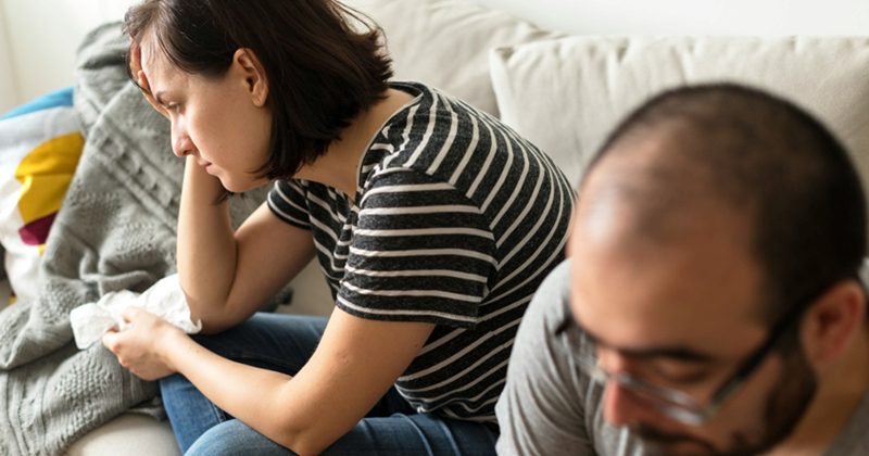A woman and a man sit on a couch together, the woman holding her head in one hand and a tissue in the other
