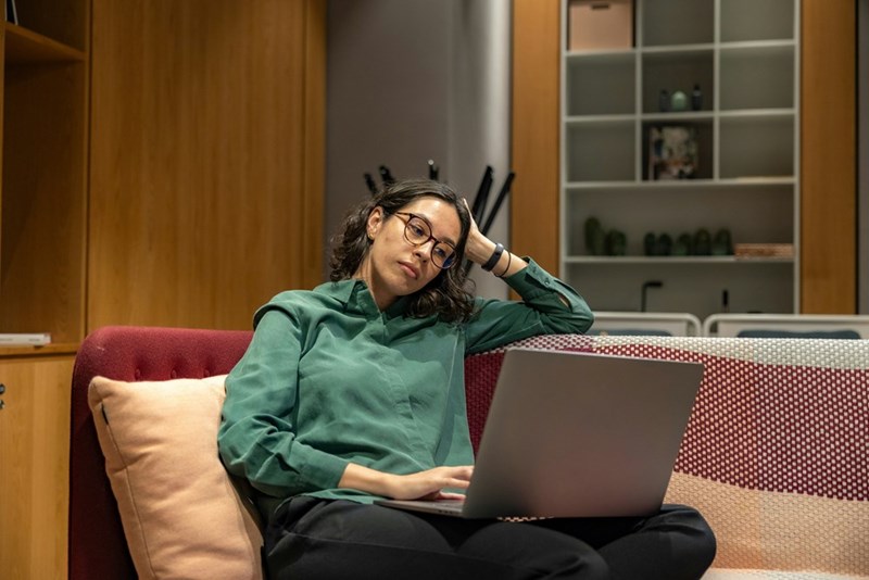 A woman sits on the couch using a laptop, looking malaiseful