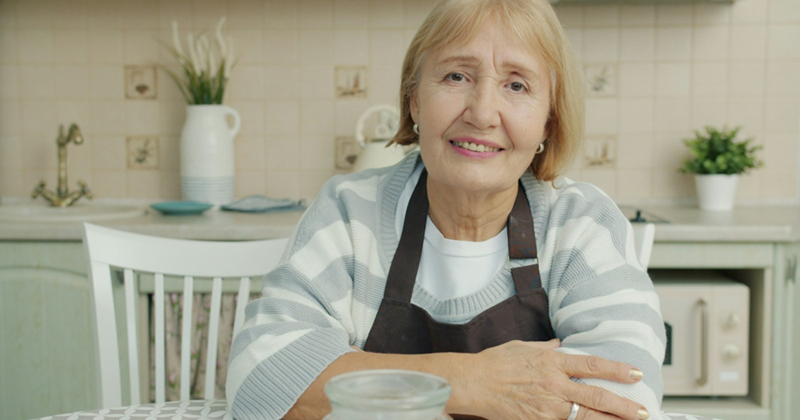 An older woman wearing an apron sits at a kitchen table