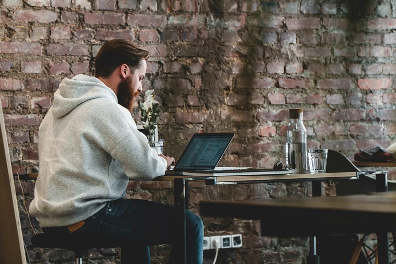 Man in hoodie working on laptop at wooden table with brick wall background.