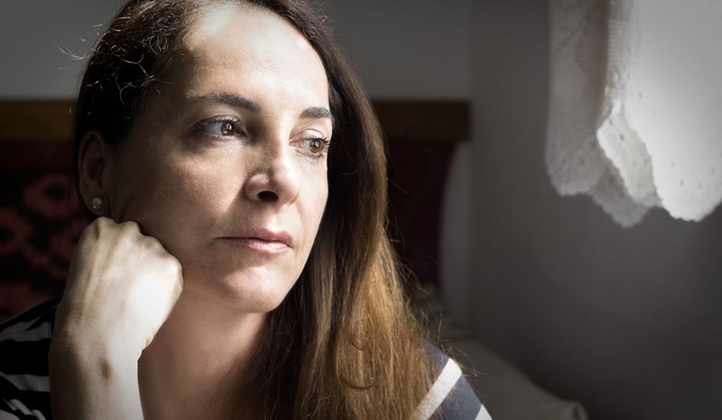Woman resting chin on hand, looking out window with pensive expression in soft natural light.
