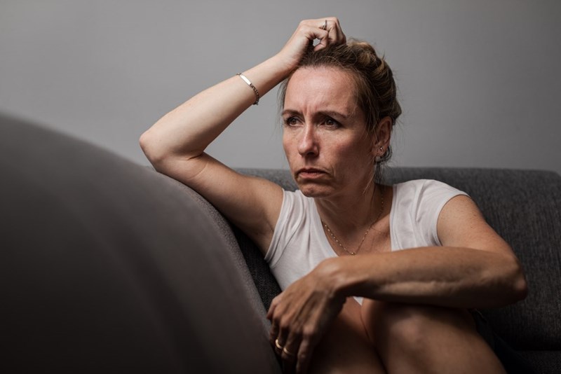 Woman sitting on couch holding her head, looking stressed and worried in indoor setting.