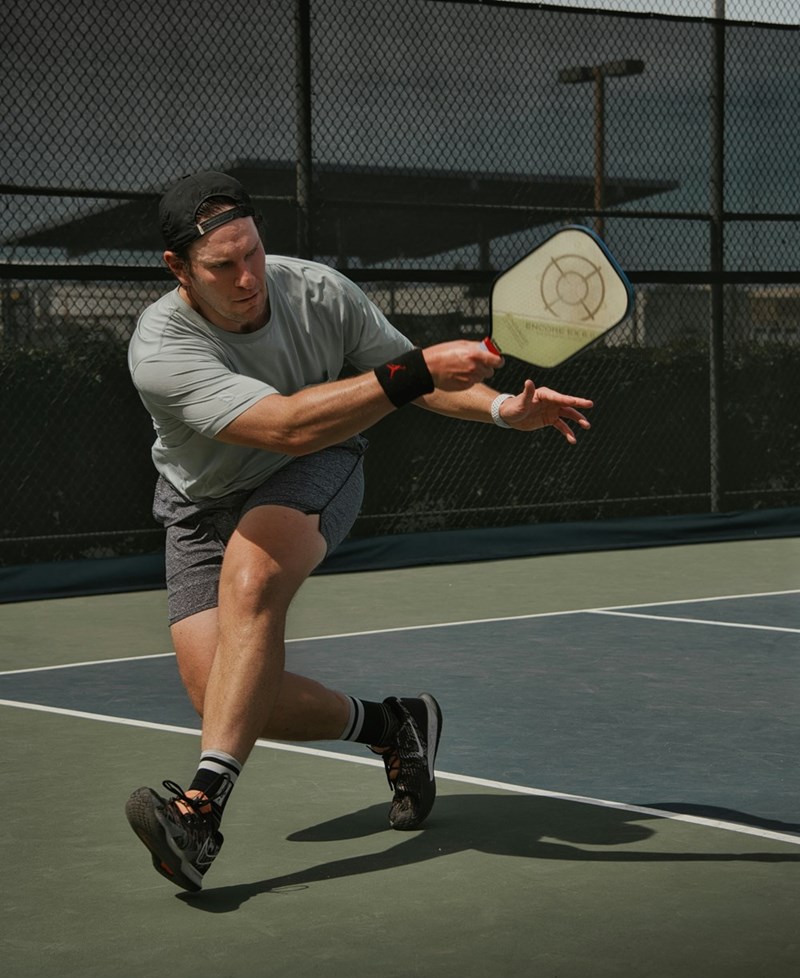 Man lunging to hit ball with paddle on outdoor court during pickleball game.