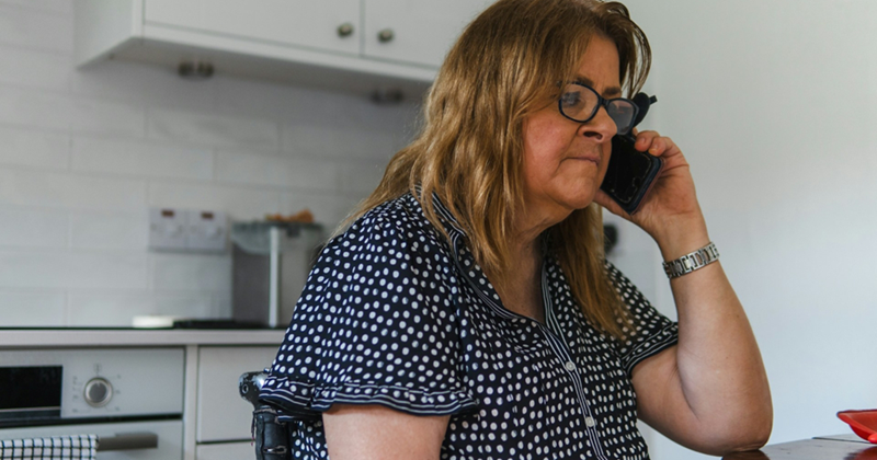 A middle-aged woman wearing glasses, talking on a cell phone while sitting at a kitchen table