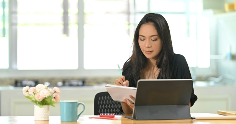 A woman working from home writes on a notepad while sitting in front of her tablet