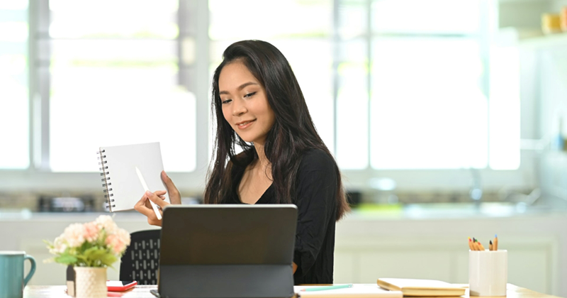 A woman working from home shows a notepad to her tablet screen as she takes a meeting
