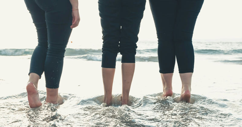 Three pairs of women's legs in rolled up dark skinny jeans, paddling in the surf at the beach