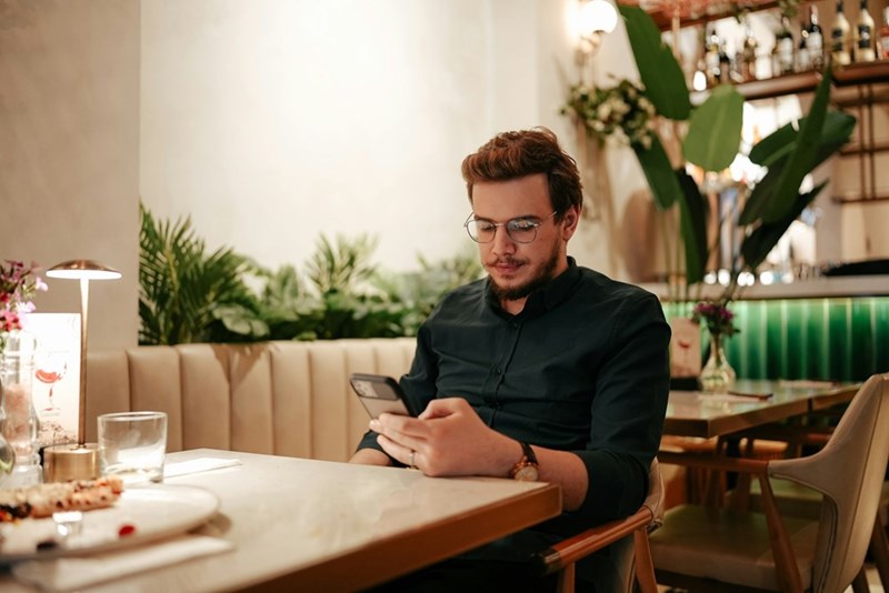 Man sitting alone at a restaurant table using his smartphone, showcasing modern dining habits and digital lifestyle in a cozy indoor setting