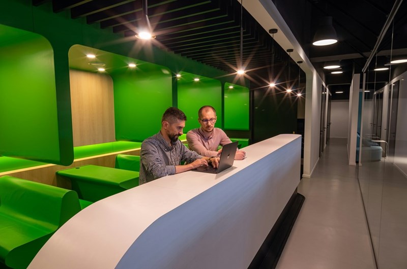 Two men collaborating on laptop at office counter with green seating in modern workspace.