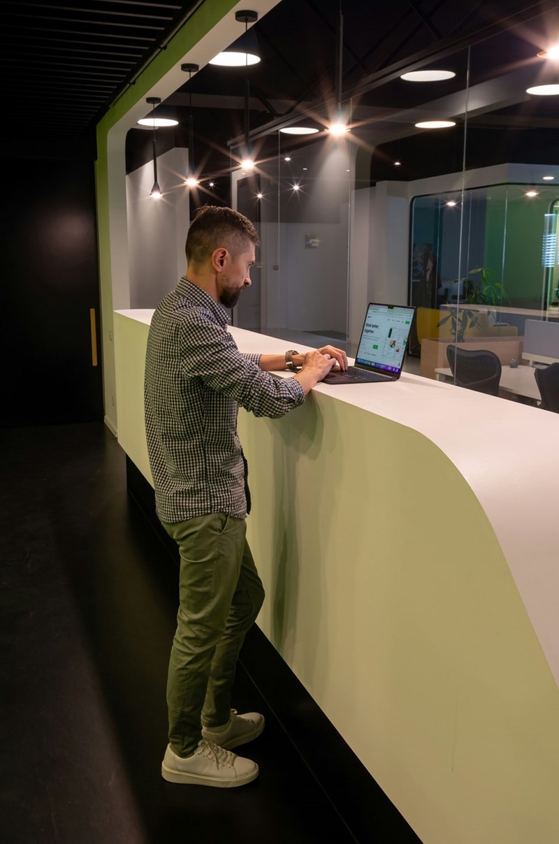 Man working on laptop at standing desk counter in modern office hallway with glass walls.