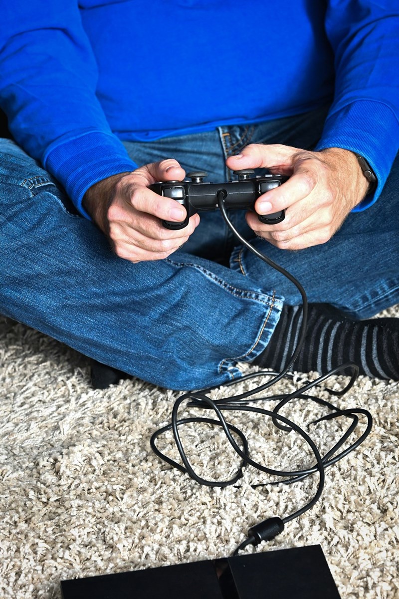 Person holding wired game controller while sitting cross-legged on carpet during video gaming session.