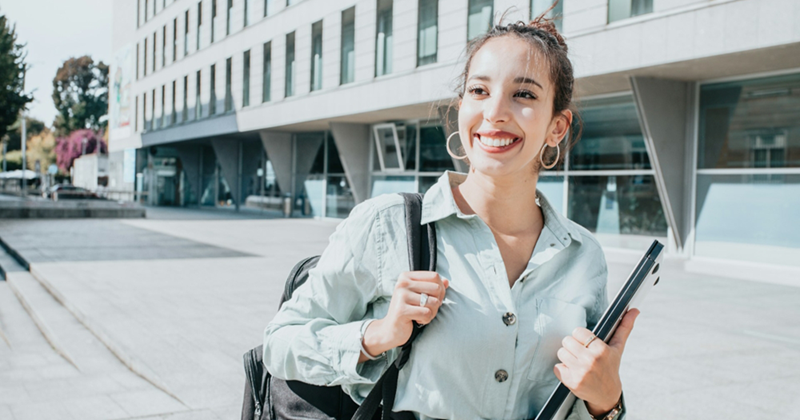 A woman smiles while walking through a college campus, holding a shoulder bag and a laptop