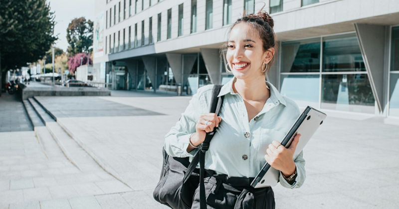 A smiling woman walks through a college campus holding a shoulder bag and a laptop