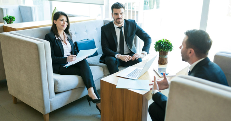 Male and female businesspeople sit on a couch opposite another businessman, who is speaking while they listen
