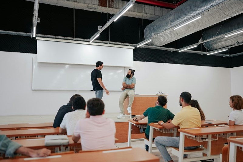 A student faces off with his professor in front of the class in a lecture hall.