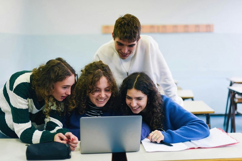 A group of unruly high school students hover around a laptop during class.