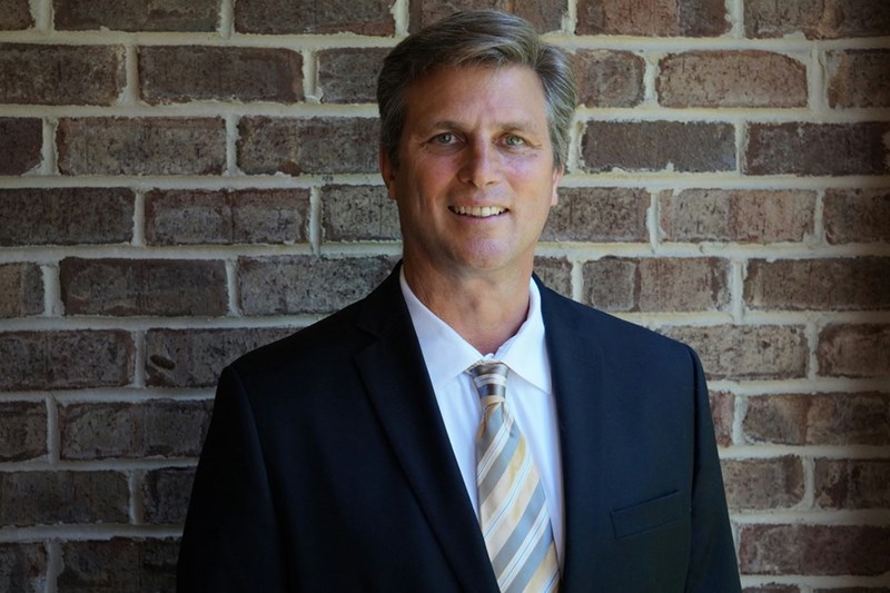 A high school principal stands against a brick wall, posing in a suit and tie.