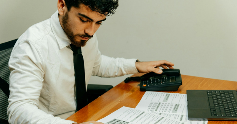 A job candidate sitting in front of a laptop has one hand on a phone, and the other writing on some papers