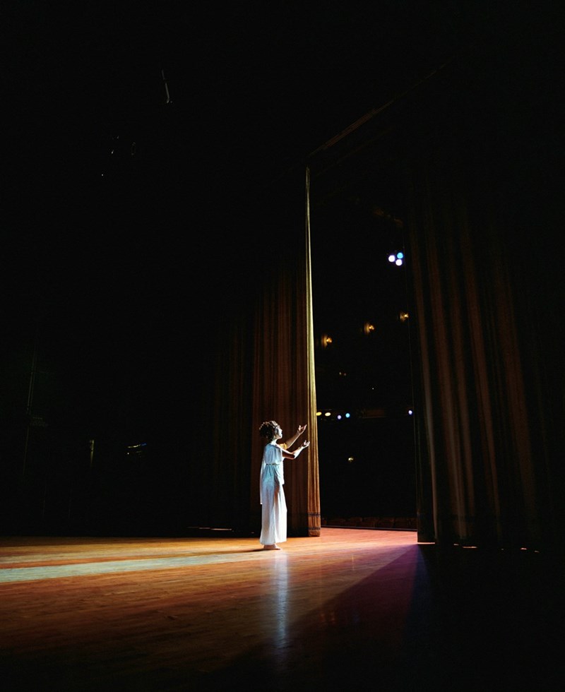 A large theater stage with red velvet curtains and an actress walking to the center