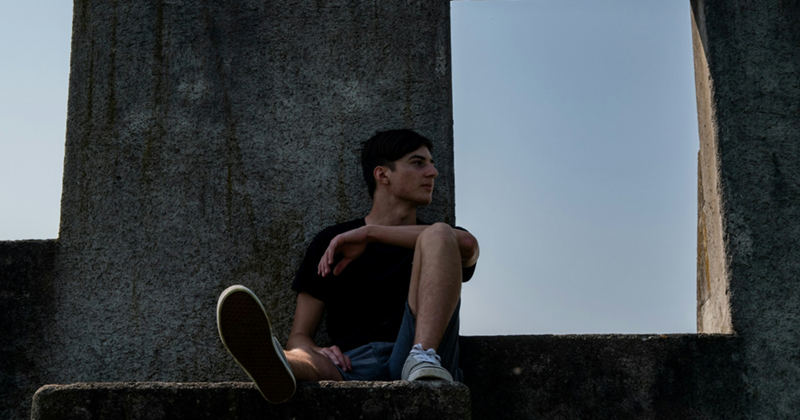 A teenage boy rests his arm on his knee and looks to the side while sitting on a concrete structure outside