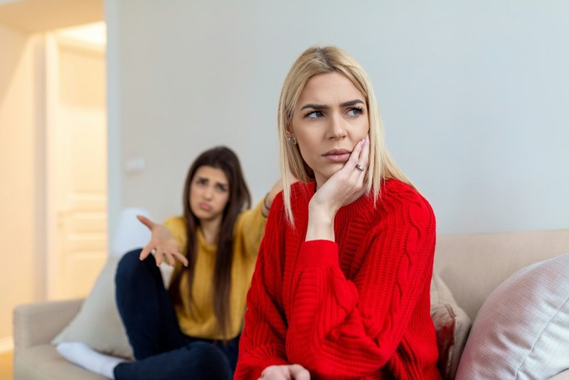 A blonde woman looks away in disappointment as her friend reaches her arms out to her while sitting on a couch.