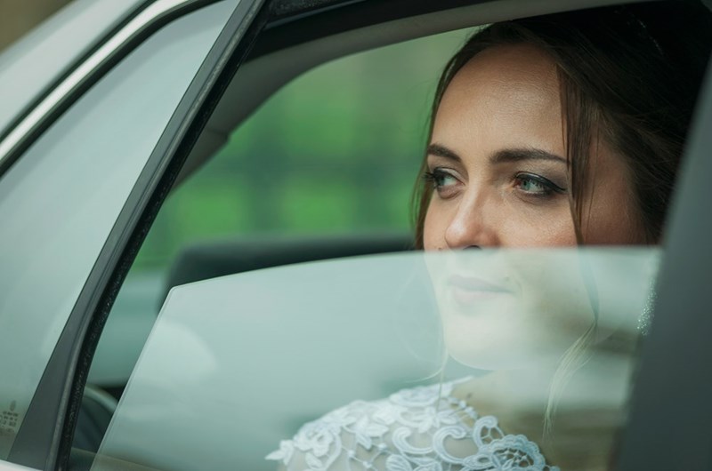 A woman in a wedding dress sits in the backseat of a car looking out the window.