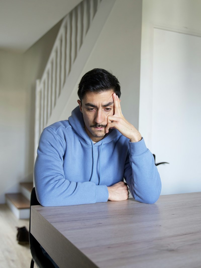 Grumpy man wearing a blue sweatshirt sits in a chair while looking at the table.